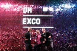Messebesuch des Vorm Teams auf der DMEXCO 2026, Pailletten-Wand in DMEXO Farben mit Leuchtmitteln in Form des DMEXCO Logos