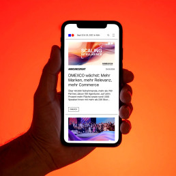 DMEXCO Webseite, Mobile Ansicht, Hand hält Telefon, Orangener Hintergrund