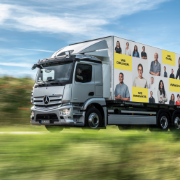 LKW f&auml;hrt durch Landschaft mit dem Visual der Mitarbeitenden Kampagne, Portraits und gelbe Textfelder mit Schrift auf LKW Plane