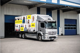 Mercedes LKW mit dem Kampagnen Motiv des Day One von Daimler Truck, Portraits und gelbe Balken mit Schrift auf Plane des LKWs