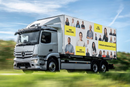 LKW fährt durch Landschaft mit dem Visual der Mitarbeitenden Kampagne, Portraits und gelbe Textfelder mit Schrift auf LKW Plane