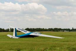 Segelflugzeug auf dem Rasen des Flugfeldes, Blaue Lackierung des Rumpfes, Slogan WinWind und Logo des Unternehmens Eraneos auf den Fl&uuml;geln