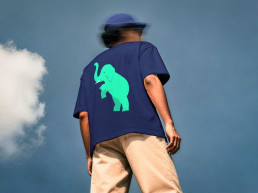 Junge Person mit Cap, stehend vor Himmel im Hintergrund, dunkelblaues Shirt mit Illustration von einem t&uuml;rkis eingef&auml;rbten Elefanten
